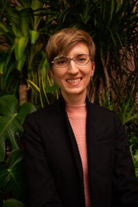 A bespectacled woman with short hair smiles in front of greenery. She wears a pink sweater and a black formal jacket. 