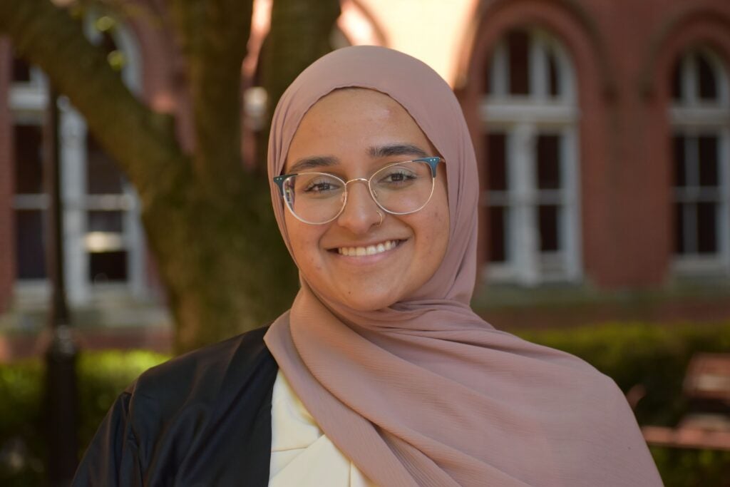 A young girl wearing glasses and a graduation gown smiles outside. She wears a pink cloth covering her hair and stands in front of an out-of-focus red brick wall. 