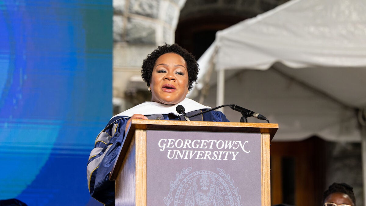 A woman with short, dark hair speaks at a wooden podium. She wears academic regalia.