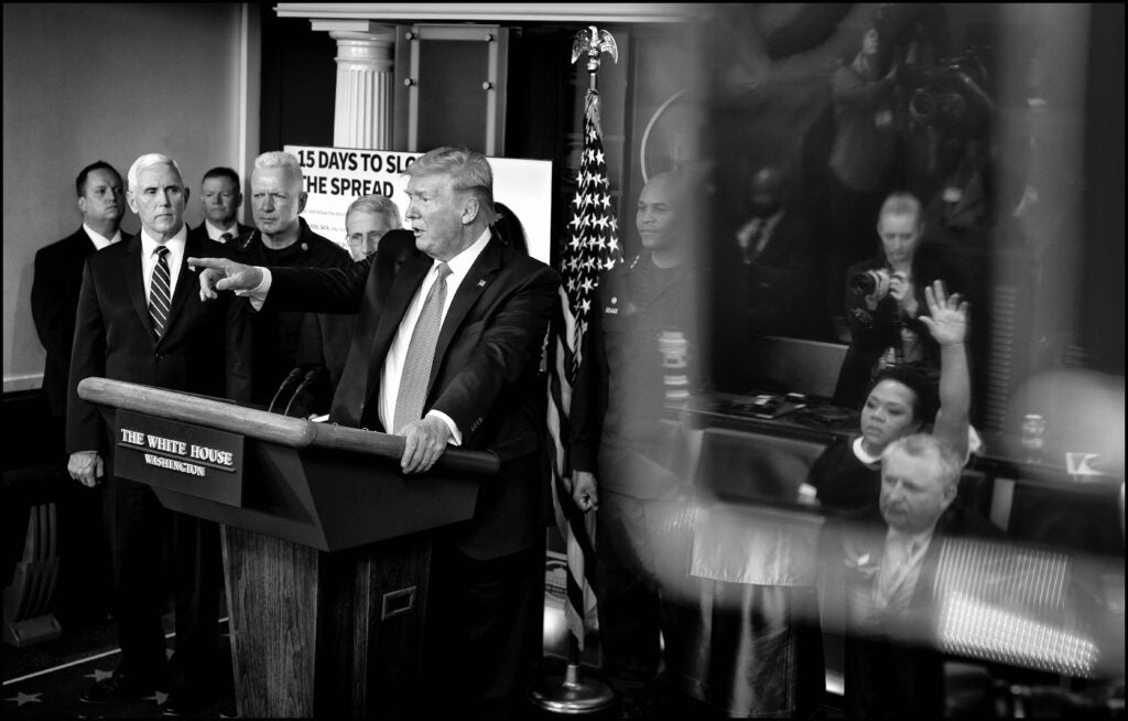 A black and white photo of a woman raising her hand to ask a question at a White House briefing. 