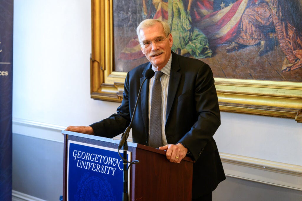 A man wearing a dark suit jacket and gray tie speaks at a podium. 