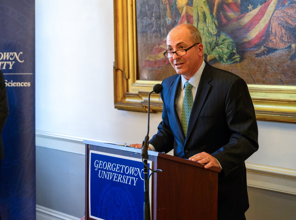 A man wearing a navy suit jacket and green tie speaks at a poodium. 