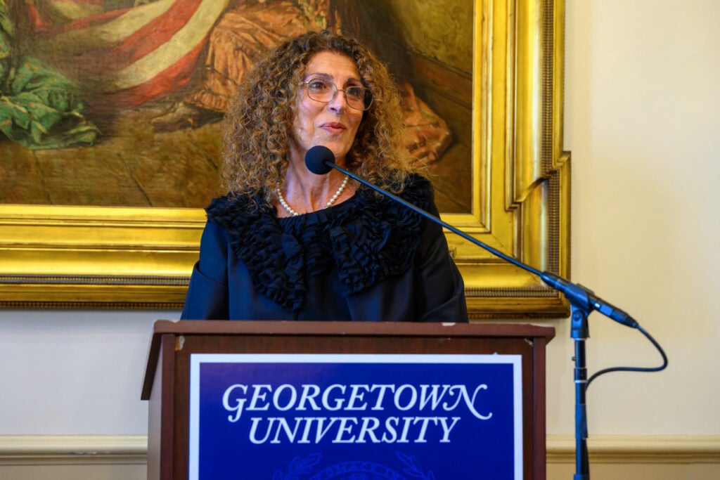 A woman with medium-length dark hair speaks at a podium. She wears a string of pearls and a formal jacket. 