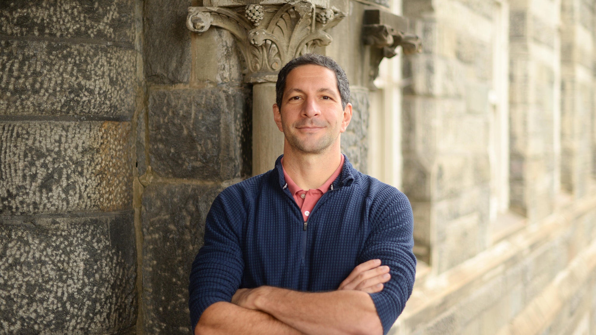 A man with short hair looks straight into the camera and crosses his arms. He stands outside and wears a blue quarterzip.