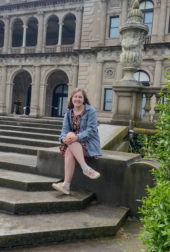 A young lady sits on stone steps out front of a large manor.