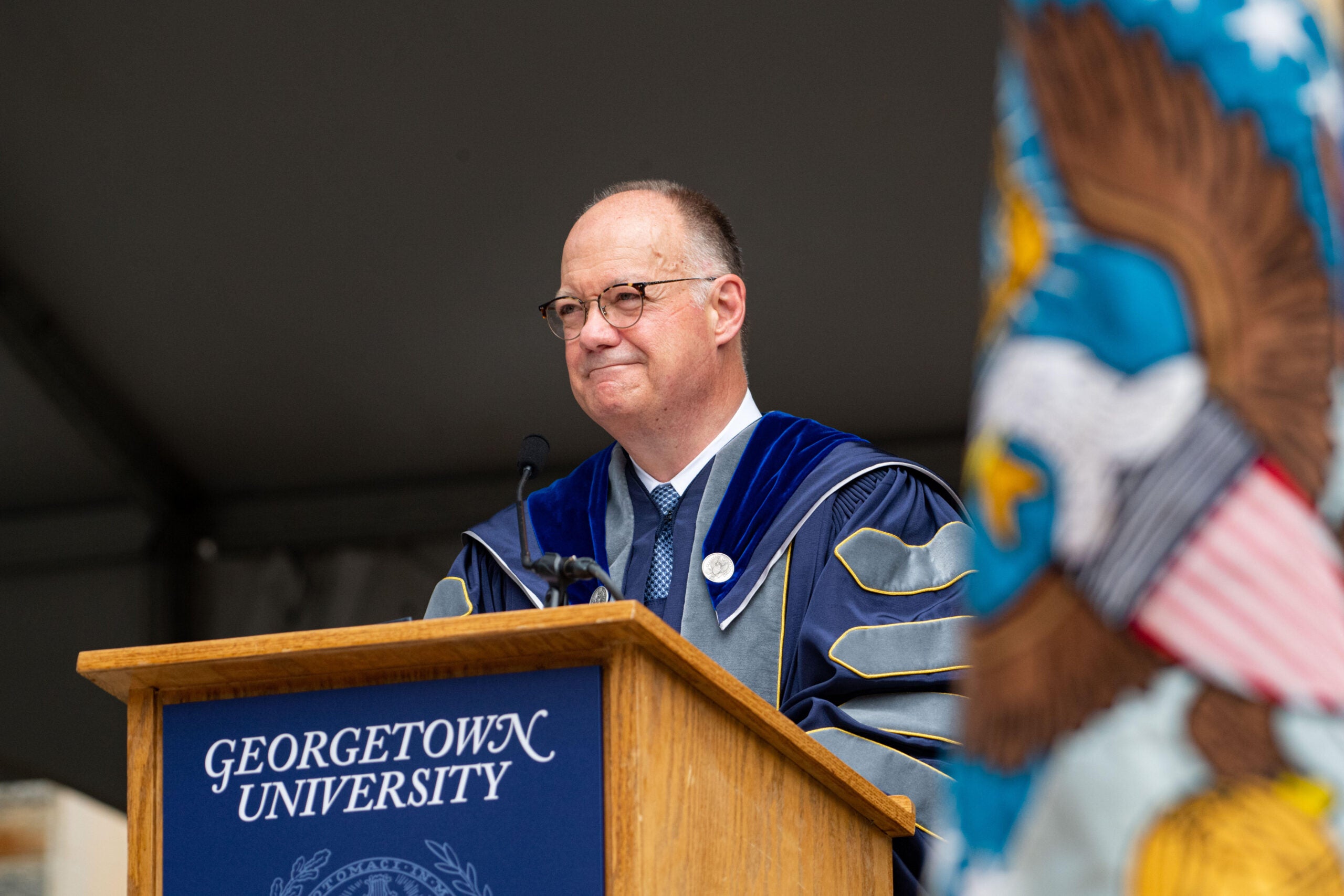 A bespectacled man in academic regalia speaks at a podium.