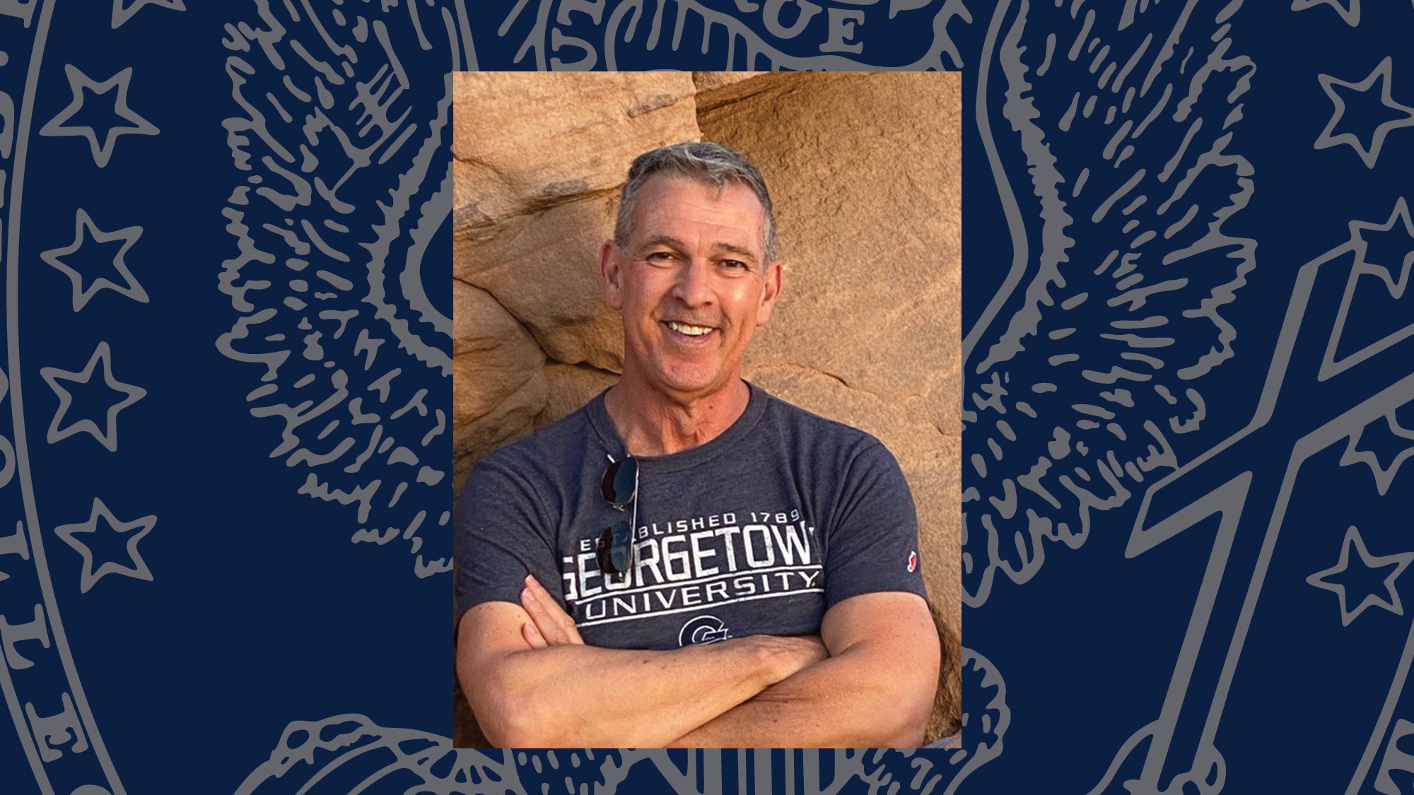 A man with short hair smiles outside. He wears at Georgetown t-shirt.