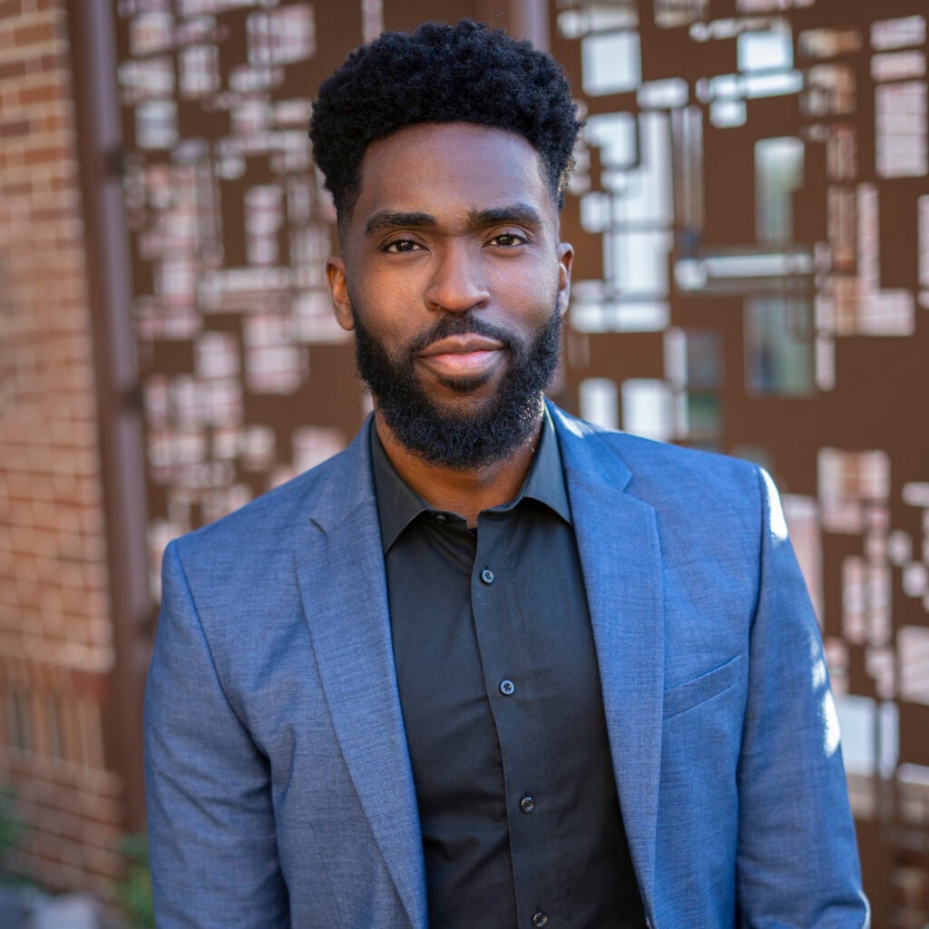 A professor wearing a black shirt with a blue jacket looking at the camera.