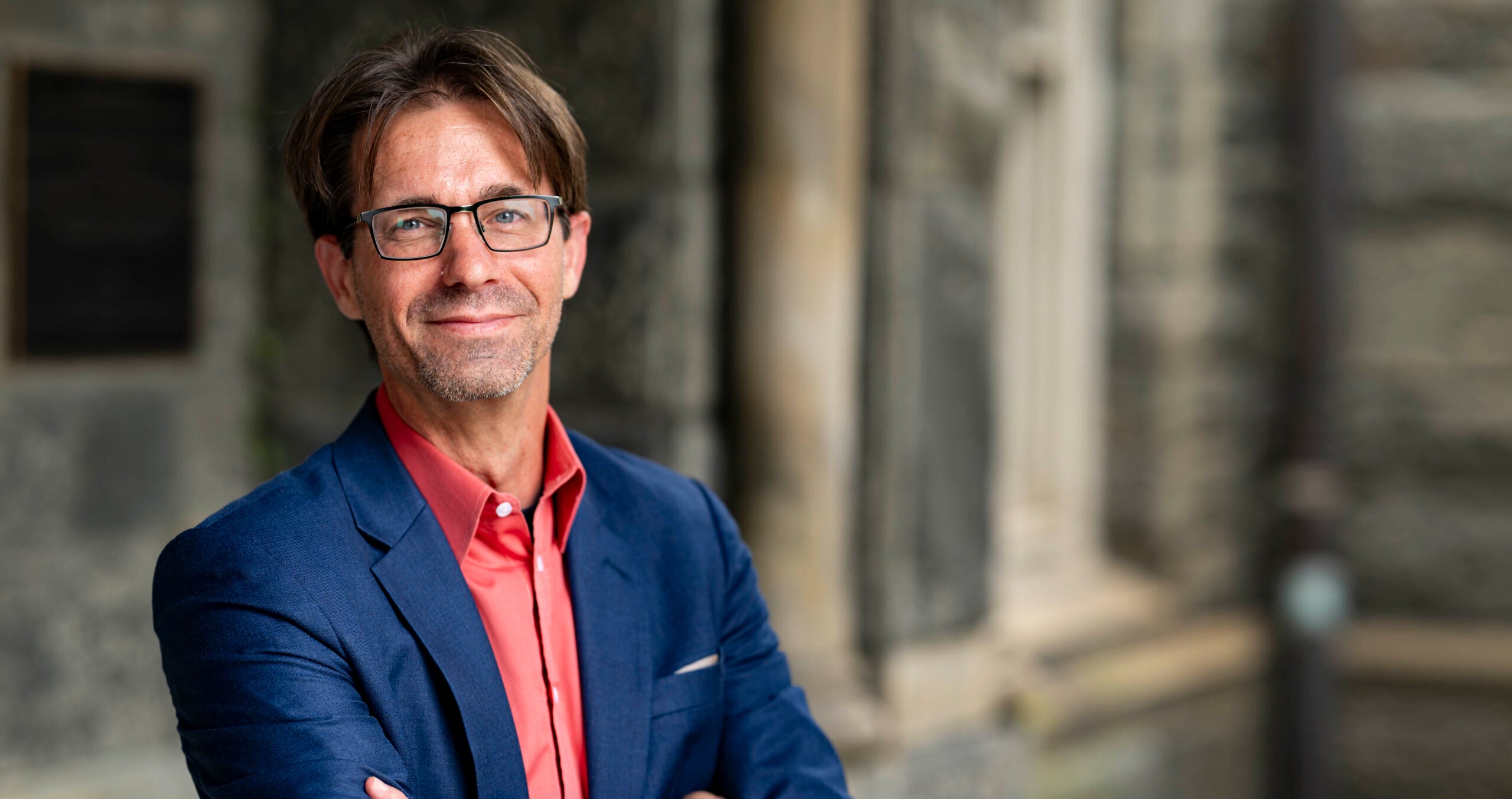 A professor wearing a blue jacket and salmon-colored shirt crossing his arms and smiling