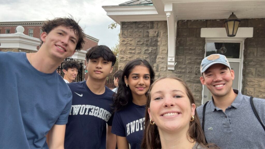 Four first-year students and a Georgetown staff member pose for a selfie in front of Georgetown University’s main gates.