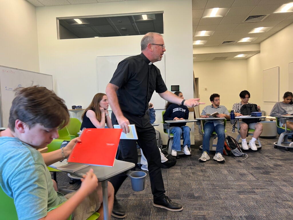 A professor stands and gestures while teaching a class, speaking to students seated around him.