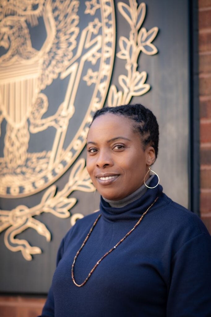 A Georgetown sociology professor wearing a blue sweater and earrings standing in front of a Georgetown University sign 