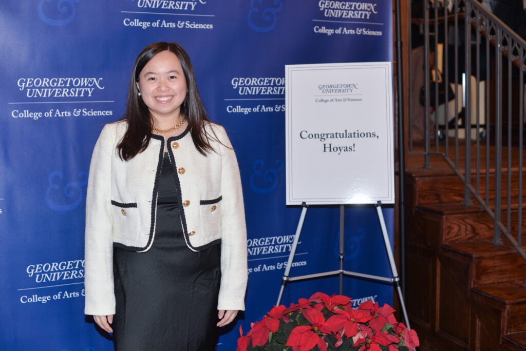 A graduating student standing in front of a Georgetown University College of Arts & Sciences banner.