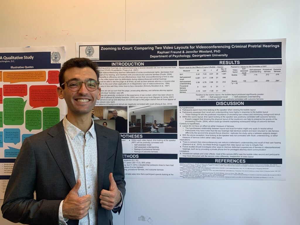 A Georgetown student wearing a shirt and jacket with his thumbs up standing in front of his thesis poster