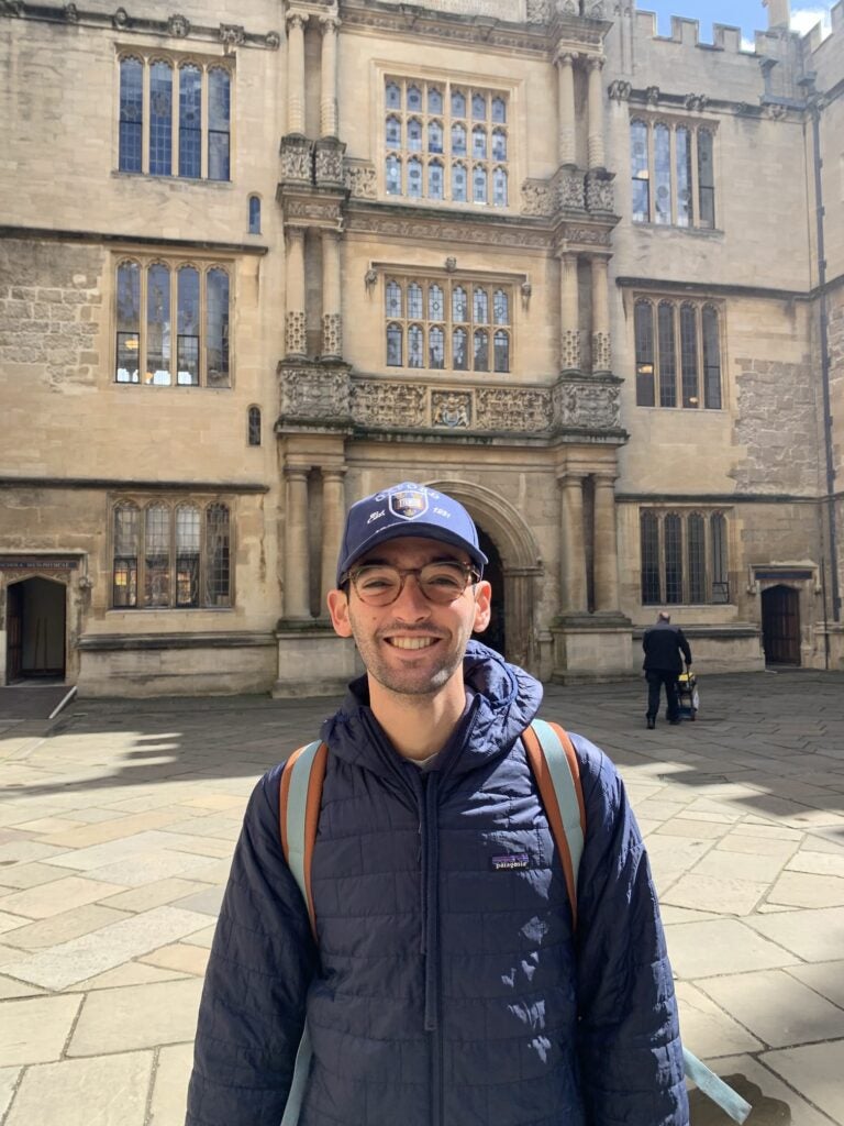 A man wearing a jacket, glasses and Oxford University hat stands in front of a building