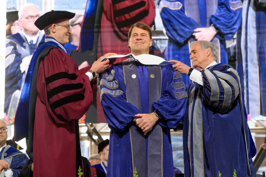A man smiles as two university leaders place an honorary degree hood on him during a commencement ceremony