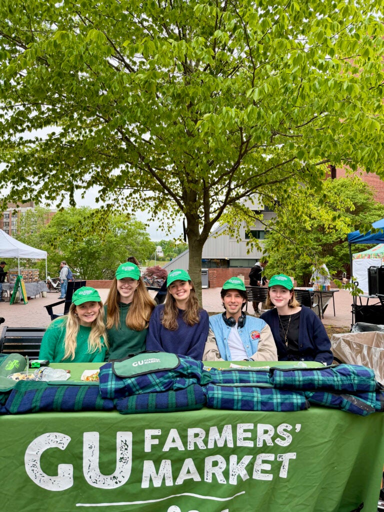 Five Georgetown students sitting behind a table that reads, GU Farmers' Market