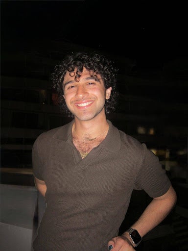 A student with short, dark curly hair smiles while standing outdoors at night.