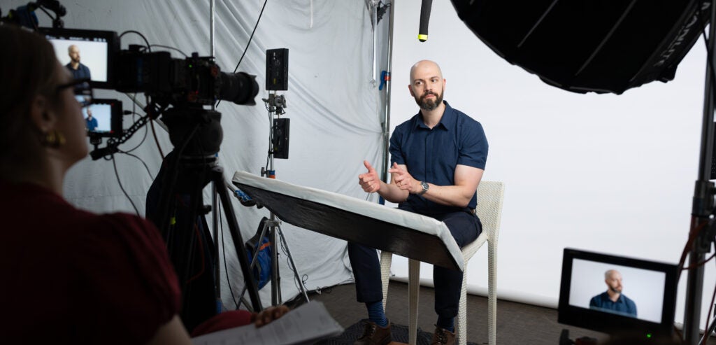 A professor wearing a blue shirt sitting in front of a camera for an interview.